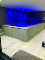 Raised indoor plunge pool glows blue under recessed ceiling lights, water still inside a wood-paneled enclosure with tiled edge, carpeted flooring and a rolled pool cover on the far wall.