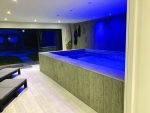 Rectangular indoor plunge pool glowing blue under recessed ceiling lights, water rippling gently; paneled wood wall with a surfboard and hanging wetsuits, sliding glass doors open to a dim patio and two loungers.