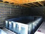 Rectangular galvanized metal pool shell sits recessed on a concrete floor, being installed inside an unfinished basement with exposed wooden joists, cinderblock walls, hanging pipes and a red tool nearby.
