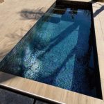 Rectangular tiled plunge pool reflects palm-tree shadows and sunlight, sitting recessed in a light stone patio with stainless edging and surrounding beige paving under a clear daytime sky.