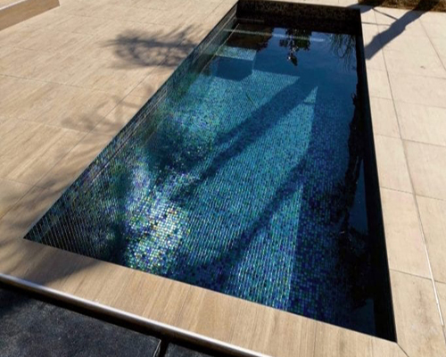 Tilestone Trainer Pool Rectangular tiled plunge pool reflects palm-tree shadows and sunlight, sitting recessed in a light stone patio with stainless edging and surrounding beige paving under a clear daytime sky.
