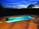 Rectangular in-ground pool glows with turquoise light, reflecting a deep blue twilight sky and silhouetted trees beyond, set within a large tiled patio at dusk.
