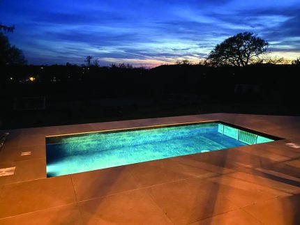 Rectangular in-ground pool glows with turquoise light, reflecting a deep blue twilight sky and silhouetted trees beyond, set within a large tiled patio at dusk.
