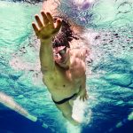 Competitive swimmer reaches forward mid stroke while propelling through turquoise lane of a pool, wearing goggles and cap, bubbles and rippling surface visible between white lane dividers.