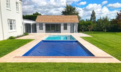 Rectangular pool partially covered by a blue solar cover, clear water at the far end, set within beige paving and green lawn before a gray pool house with glass doors.