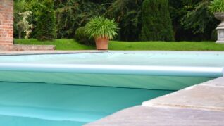 AQM-Aquaeous-Inwall-Sage-Green-Slice-1 A turquoise pool cover rolls partially across a backyard swimming pool, with stone coping at the edge, brick wall, green lawn, potted plants and an ornamental urn beyond.
