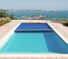 Rectangular pool sits filled with clear turquoise water, partially covered by a blue solar cover, surrounding tiled terrace overlooks a glass-railed coastal vista with a sandy peninsula and distant town.