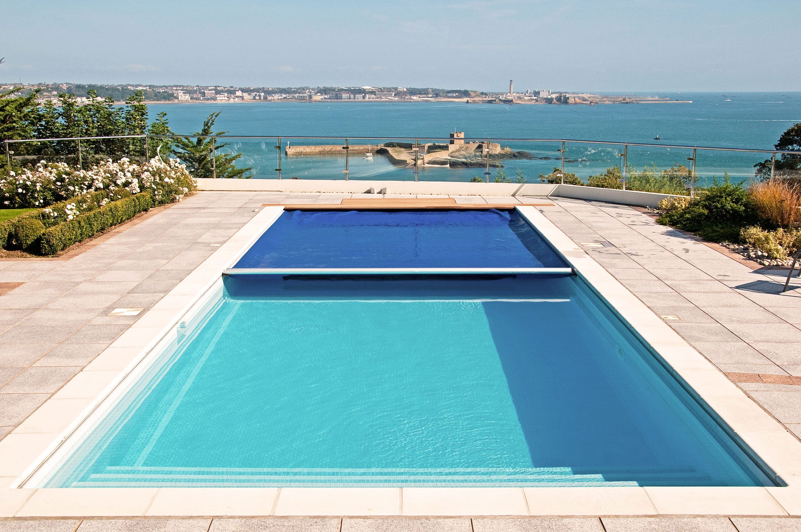 Rectangular pool sits filled with clear turquoise water, partially covered by a blue solar cover, surrounding tiled terrace overlooks a glass-railed coastal vista with a sandy peninsula and distant town.