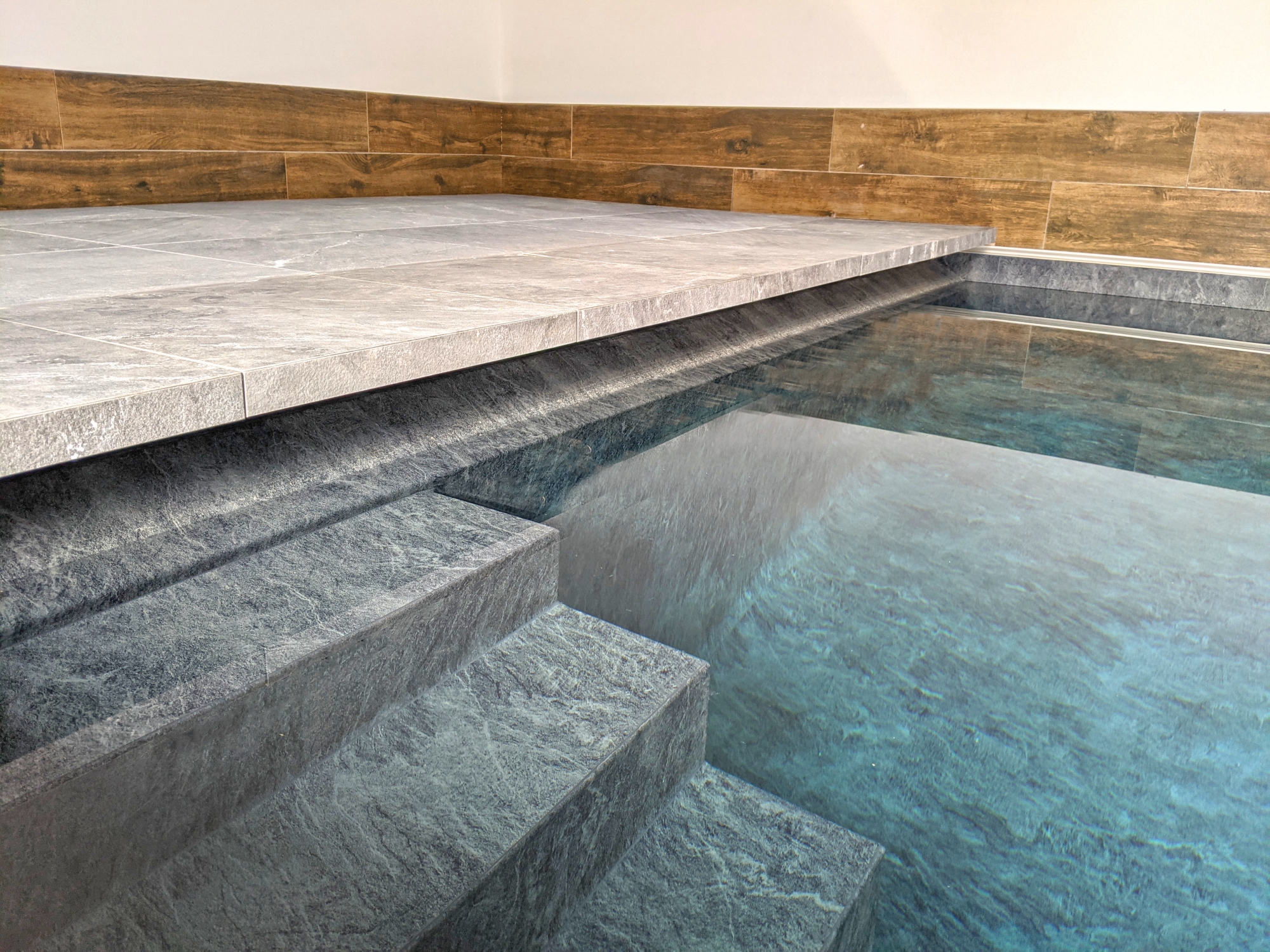 Stone pool steps descend into clear blue water while a raised tiled platform overhangs above, surrounded by wood look wall tiles and white walls indicating an indoor pool area.
