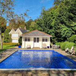 AQM Inwall Royal Blue 1 A rectangular outdoor swimming pool covered with a blue cover lies centered on a stone patio, facing a small cream pool house; lawn, sunlit trees and lounge chairs surround it.