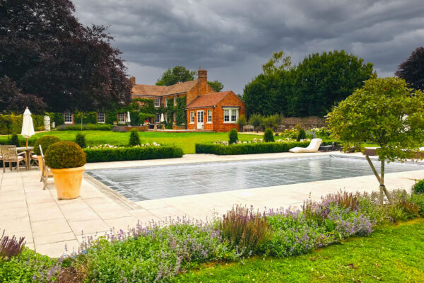 A covered rectangular swimming pool reflects a dark stormy sky, sitting on a paved terrace surrounded by potted shrubs, lounge chairs, flowerbeds and a red-brick country house amid lawns.