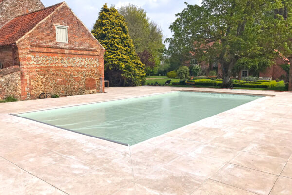 Outdoor rectangular swimming pool sits covered with a pale green cover, surrounded by light stone decking, adjacent to an old red-brick barn and a landscaped garden with trees and hedges.
