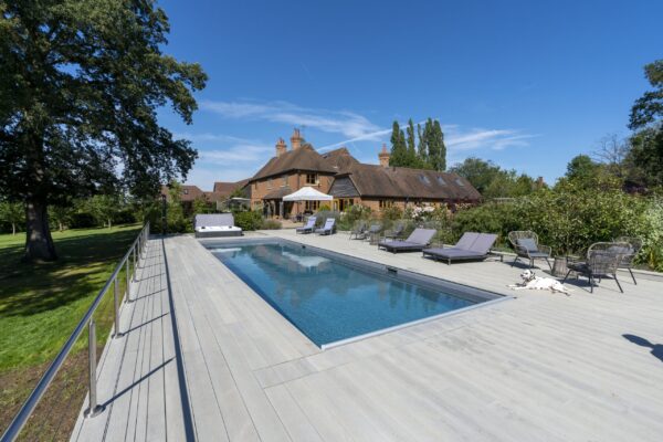 Rectangular swimming pool reflects blue sky, bordered by pale deck; loungers and a spotted dog lie beside it, with a large brick house and trees in the sunny garden.