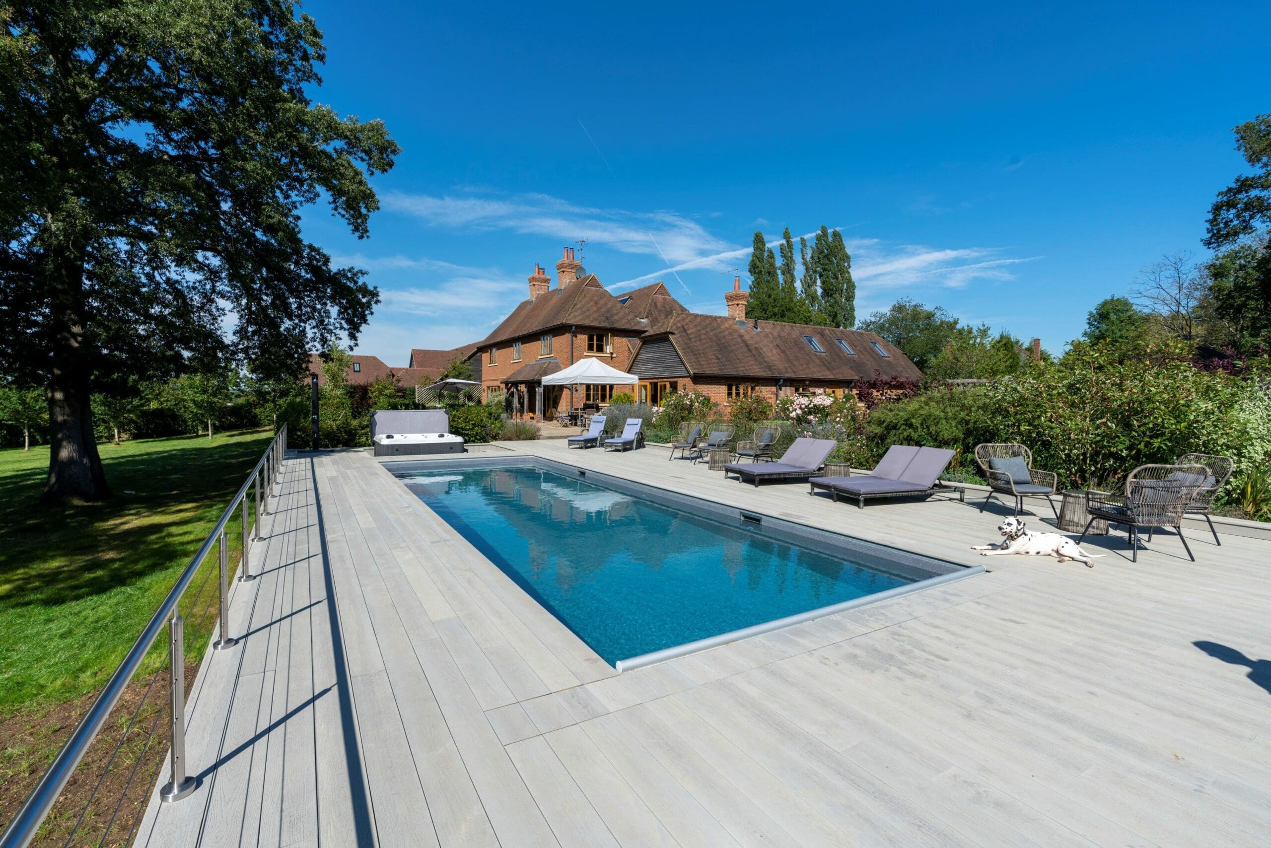 Rectangular swimming pool sits within a wide pale wood deck, reflecting the blue sky; lounge chairs and a Dalmatian recline nearby, with a large brick house and trees in the background.