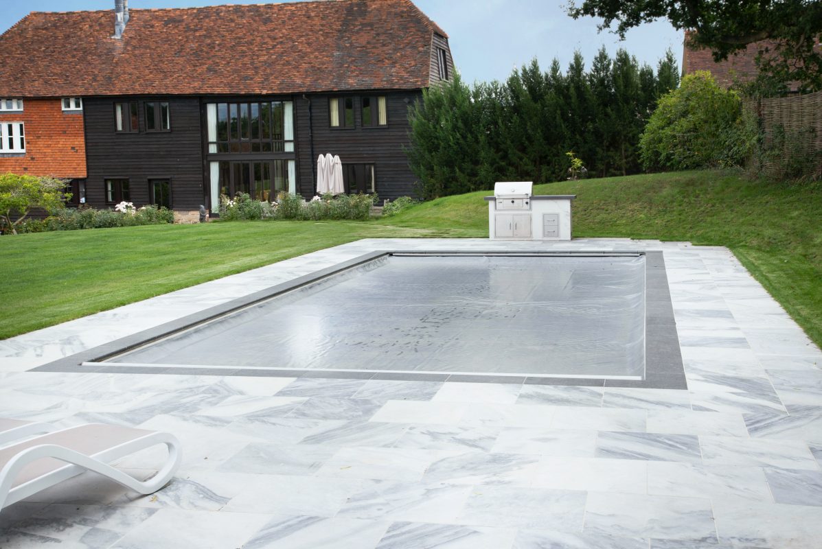 AQM-Mermaid_Pools_Inwall-track-light-grey-fabric A rectangular pool is covered with a taut safety cover, set within white marble paving beside green lawn, an outdoor grill and a dark timber house with red tiled roof.