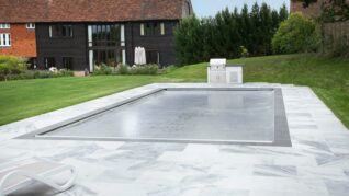 AQM-Mermaid_Pools_Inwall-track-light-grey-fabric A rectangular pool is covered with a taut safety cover, set within white marble paving beside green lawn, an outdoor grill and a dark timber house with red tiled roof.