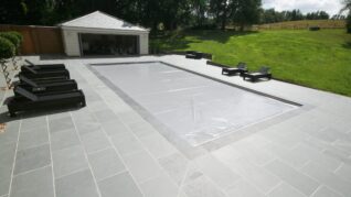 AQM-Tanby-Inwall-Light-Grey- Rectangular in-ground pool covered with a gray protective sheet, surrounded by tiled patio with black lounge chairs, an open pool house at rear and a grassy slope with trees.