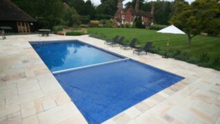 Navy Blue AQM Pool Cover A rectangular swimming pool partially covered by a blue solar cover, set on a pale stone patio with lounge chairs and umbrella beside a large lawn and brick house.