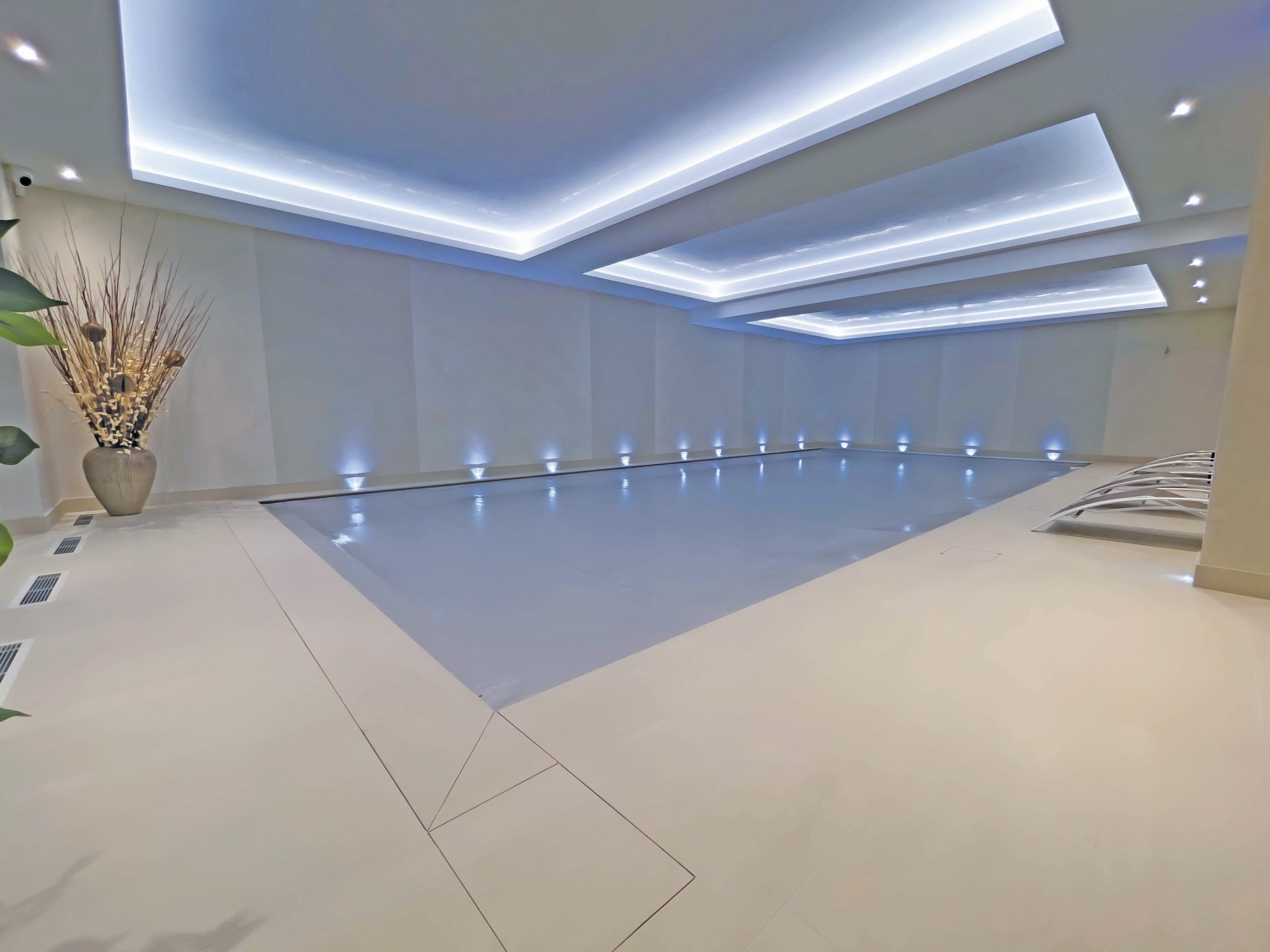 Indoor rectangular swimming pool sits covered in a minimalist spa room with recessed LED ceiling panels, blue uplights along walls, beige deck, vase of dried branches and loungers.