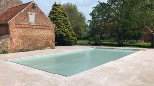 AQM-sage-green-Inwall-2 Rectangular outdoor pool covered with a taut pale blue safety cover, set within a wide stone patio, adjacent to an old brick barn and mature trees bordering a manicured lawn.