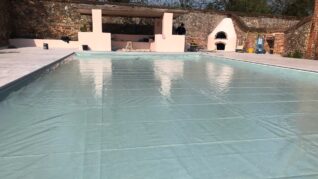 AQM-sage-green-Inwall-4 An empty pool is being fitted with a pale turquoise liner, its glossy surface reflecting a pink pavilion and a worker, set within a sunlit stone deck and brick courtyard.
