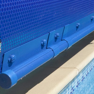 Blue pool cover roller with interlocking slats secured by plastic hinges sits along beige pool coping above blue mosaic tiled water, partially rolled and mounted at the pool's edge.