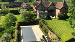 Charcoal Grey AQM 2 Brick country house sits beside a manicured lawn and a covered rectangular swimming pool, with patio furniture, hedged gardens, trees and neighboring outbuildings set amid rolling green fields.