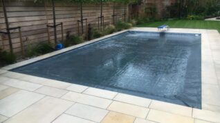 Charcoal Grey AQM 3 Rectangular inground swimming pool covered by taut dark safety cover, set in pale stone patio with a blue diving board, lawn, young trees and a wooden privacy fence.