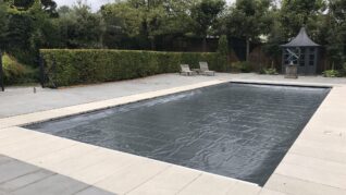 Charcoal Grey AQM Rectangular outdoor pool sits covered by a dark protective sheet, surrounded by pale stone paving, two loungers, a small gazebo, trimmed hedges and tall trees under a cloudy sky.