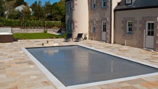 Charcoal Grey AQM 4 Rectangular in-ground pool sits covered with a taut gray safety cover, surrounded by light stone patio beside a beige two-story house with doors and lounge chairs, lawn and trees beyond.