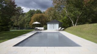 Dark Grey AQM 2 Rectangular pool covered by a tarp sits on a paved terrace with lounge chairs, umbrella, and blue poolhouse, surrounded by lawn and tall trees under a cloudy sky.
