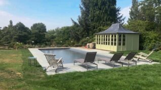 Dark Grey AQM 3 Rectangular swimming pool sits on a stone patio in a grassy backyard, bordered by four gray lounge chairs and two deck chairs, with a pale green pavilion and tall trees beyond.