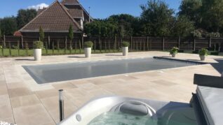Dark Grey AQM A covered rectangular pool sits on a pale tiled patio; a bubbling hot tub fills the foreground, with potted shrubs, a wooden fence and a house with tiled roofs beyond.