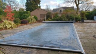 Dark Grey AQM 4 Rectangular swimming pool covered with a translucent plastic pool cover stretched within metal rails, set on a flagstone patio surrounded by trimmed shrubs, potted plants, leaf-strewn ground and houses.
