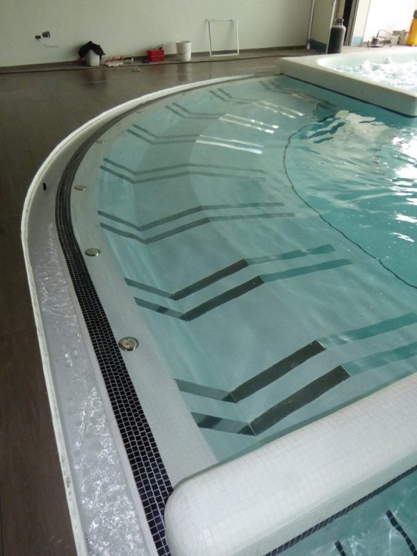 Double Air Bed - 270mm wide - Concrete Curved indoor pool steps, water rippling over tiled risers with chevron stripes while an adjacent raised spa bubbles, tiled overflow channel and dark wood deck with tools against far wall