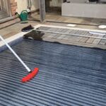 Pool cover being brushed with a red broom on a residential patio beside stone tiles and a wooden deck, near a green watering can and house steps.