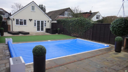 Light Blue AQM 3 Rectangular in-ground pool is covered by a blue tarp in a paved suburban backyard with a cream bungalow, patio furniture, potted spherical topiary and an ivy-covered dark fence © www.funfantastic.com