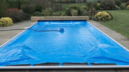 Light Blue AQM Rectangular outdoor swimming pool covered by a blue solar cover with a small pump and hose resting on it, set on a stone patio surrounded by shrubs, lawn, trees and distant hills.