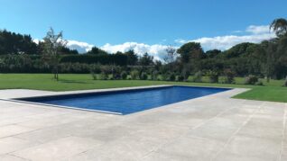 Navy Blue AQM Pool Cover Rectangular in-ground swimming pool sits covered with a taut blue safety cover, recessed in a pale stone patio, surrounded by trimmed lawn, scattered trees and hedges beneath a blue sky.