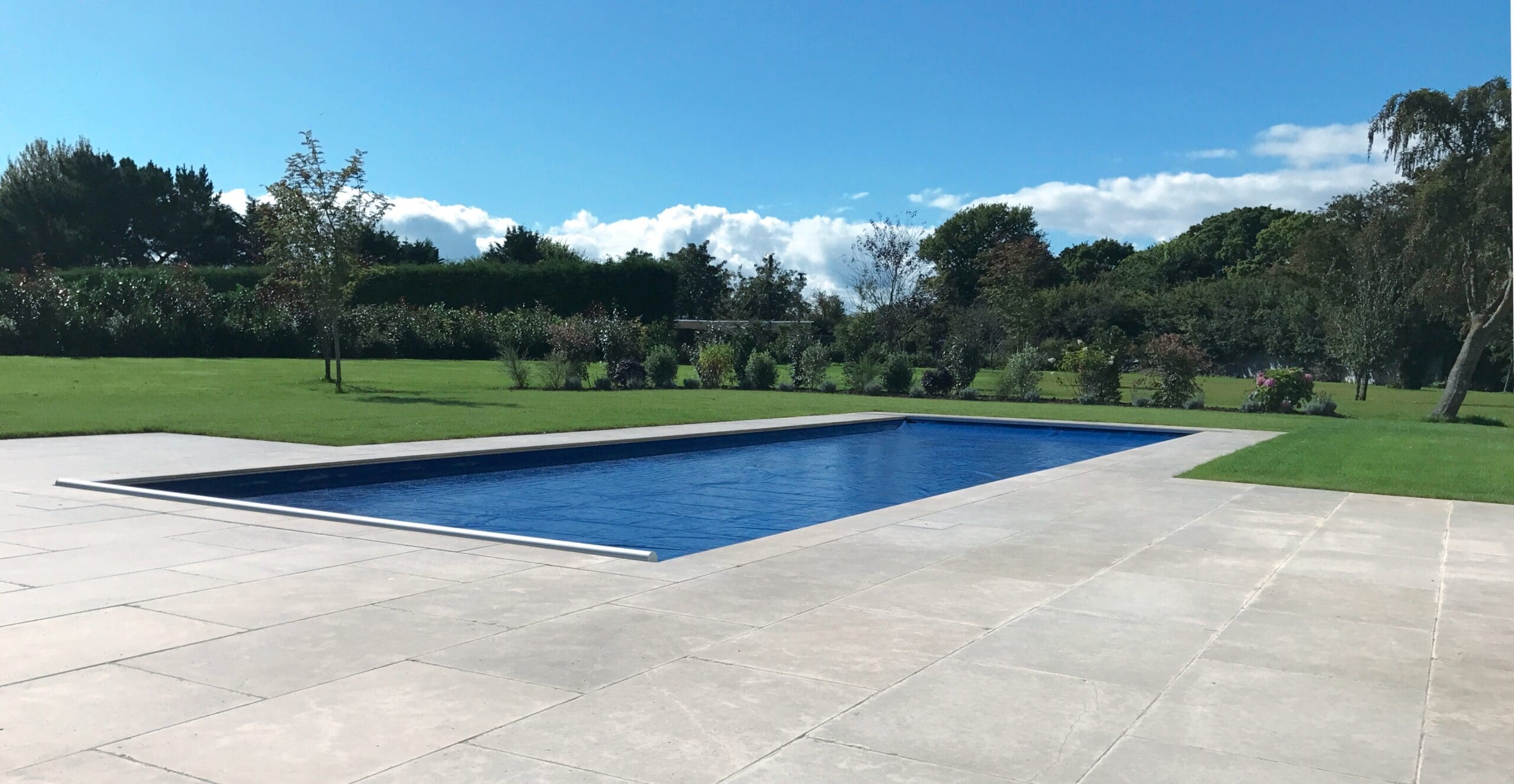 Rectangular in-ground swimming pool sits covered with a taut blue safety cover, recessed in a pale stone patio, surrounded by trimmed lawn, scattered trees and hedges beneath a blue sky.