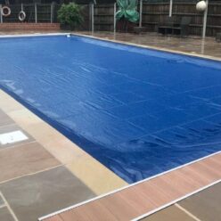 Royal Blue AQM 2 Rectangular outdoor swimming pool covered by a blue solar cover, sitting on a wet stone terrace with metal ladder, life rings and wooden fence in the background.