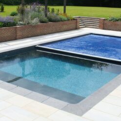Royal Blue AQM 3 Rectangular swimming pool partially covered by a blue solar cover, reflecting sunlight and gentle ripples; set in pale tiled patio with brick retaining wall, flowerbeds and a manicured lawn, chaise lounges at right.