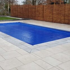 Royal Blue AQM 4 Rectangular in-ground pool covered by a taut blue cover, set within pale stone coping, surrounded by light tiled patio and tall wooden slatted fence with lawn and bare trees beyond.