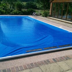 Royal Blue AQM Rectangular swimming pool covered by a blue solar cover, slightly wrinkled and secured at one end, set beside a brick house with glass doors and a surrounding patio and garden.
