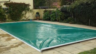 Sage Green AQM 2 Rectangular outdoor pool covered by a green plastic tarp with wrinkles; it sits on a stone-tiled patio flanked by potted plants, climbing vines on a pale wall and dense hedges.