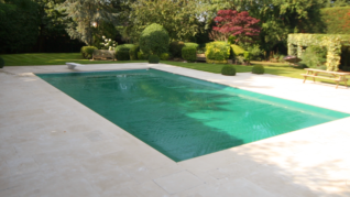 Sage Green AQM 3 Rectangular turquoise pool ripples gently, set in a pale stone patio, framed by trimmed hedges, shrubs and trees with a bench, picnic table and diving board in a sunny garden.