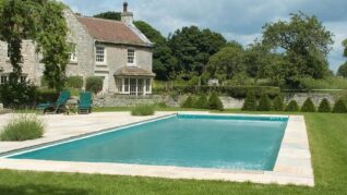 Sage Green AQM A rectangular turquoise swimming pool sits calm on a lawn, bordered by stone paving; two green lounge chairs face the water while a stone house and trees fill the background.