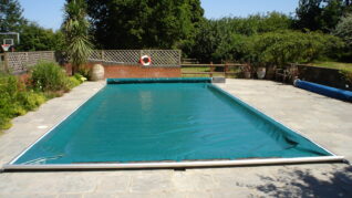 Sage Green AQM 4 Rectangular outdoor pool covered by a teal safety cover rolled at far end; stone patio surrounds it, with potted plants, a fence and trees in the garden beyond.