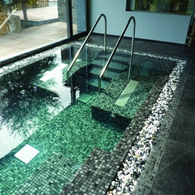 Single Air Bed 150mm wide - Concrete A small indoor mosaic-tiled plunge pool with metal handrails, stepped seating descends into clear green water, light reflecting on tiles, pebble border and slate floor beside large glass windows.