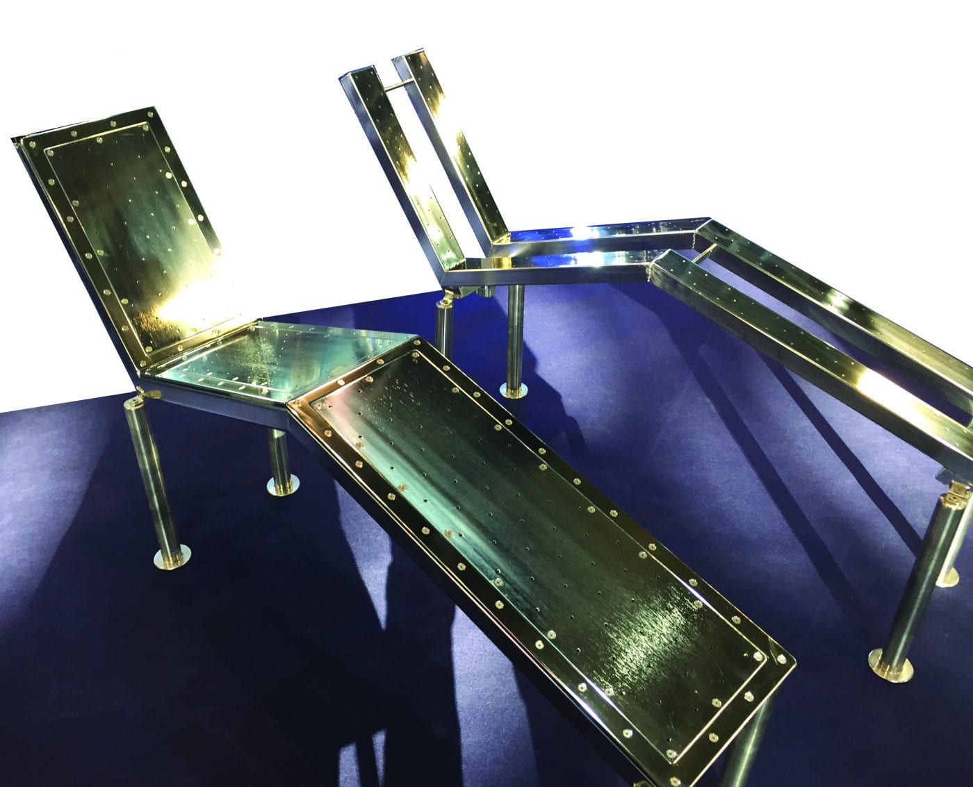 Single Air Bed – 210mm wide – Liner Polished brass-and-black metal lounge chairs extend and recline on a deep blue carpet, panels riveted and reflecting bright light, casting long angular shadows across the floor.